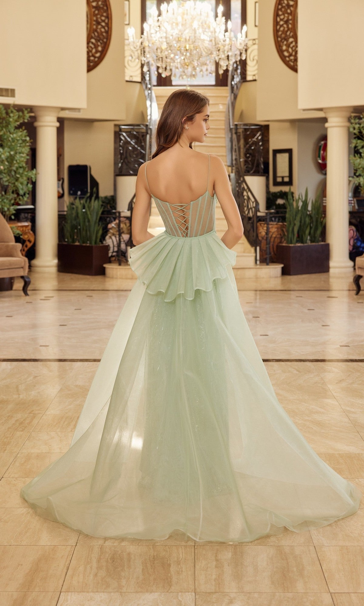A woman wearing the Nox Anabel N1763 long prom dress in mint green, featuring a floor-length silhouette and layered ruffled back, stands indoors facing away amid columns, plants, and a chandelier.